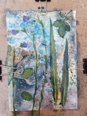 Artistic paper with green leaves and blue watercolor patterns, held together by clips on a wooden surface.