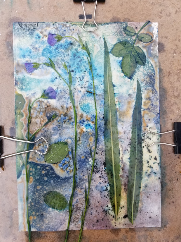 Artistic paper with green leaves and blue watercolor patterns, held together by clips on a wooden surface.