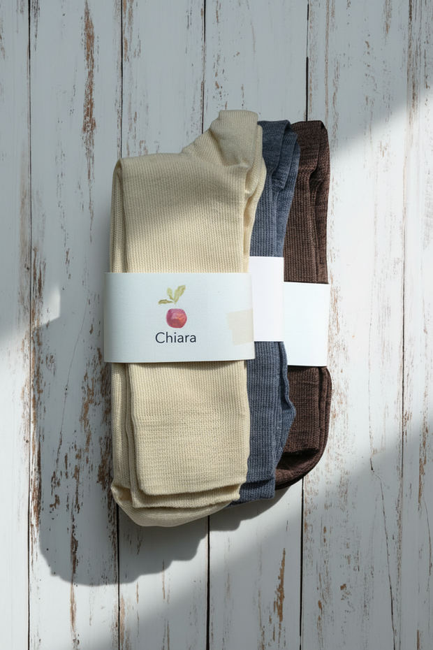3 pairs of white, grey and brown wool socks on a white rustic wooden background with a white label showing Chiara and a red apple