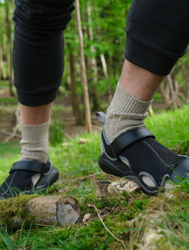 Person wearing black sandals and beige socks standing on a grassy area with greenery in the background