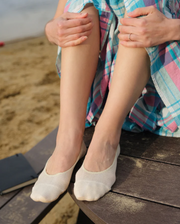 Person sitting on a wooden bench by the beach wearing white socks and a colorful dress.