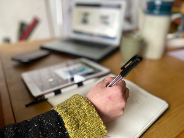 Person holding a pen over a notebook with a blurred background of a desk with a laptop and mug.