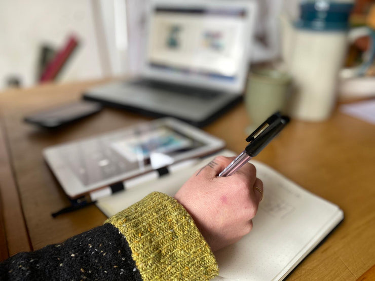 Person holding a pen over a notebook with a blurred background of a desk with a laptop and mug.