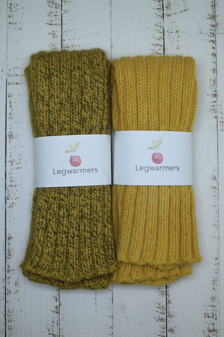 Two pairs of knitted yellow legwarmers on a wooden surface with labels.